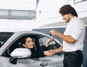 A car dealer hands over keys to a happy woman seated in her new car. They shake hands to finalize the deal.