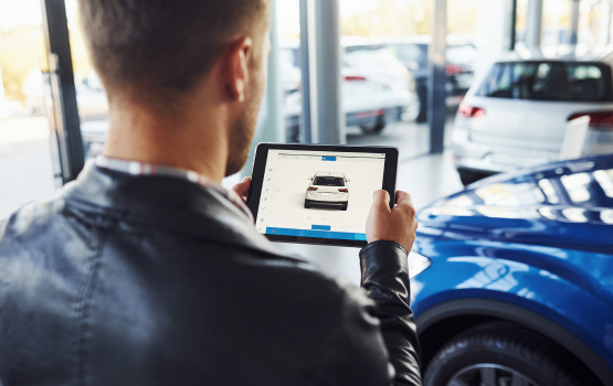 A man in a leather jacket holds a tablet displaying a car listing inside a modern dealership with vehicles in the background.