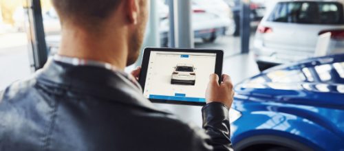 A man in a leather jacket holds a tablet displaying a car listing inside a modern dealership with vehicles in the background.