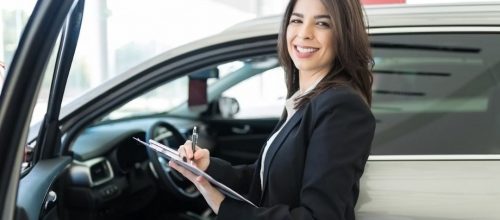 A smiling salesperson in a black suit stands beside a car in a showroom, taking notes on vehicle inventory. This image reflects the importance of optimizing car inventory management to drive profit.