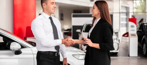 A man and woman shake hands in a car dealership showroom, finalizing a deal. Automotive Inventory Management Partner can help streamline your sales process.
