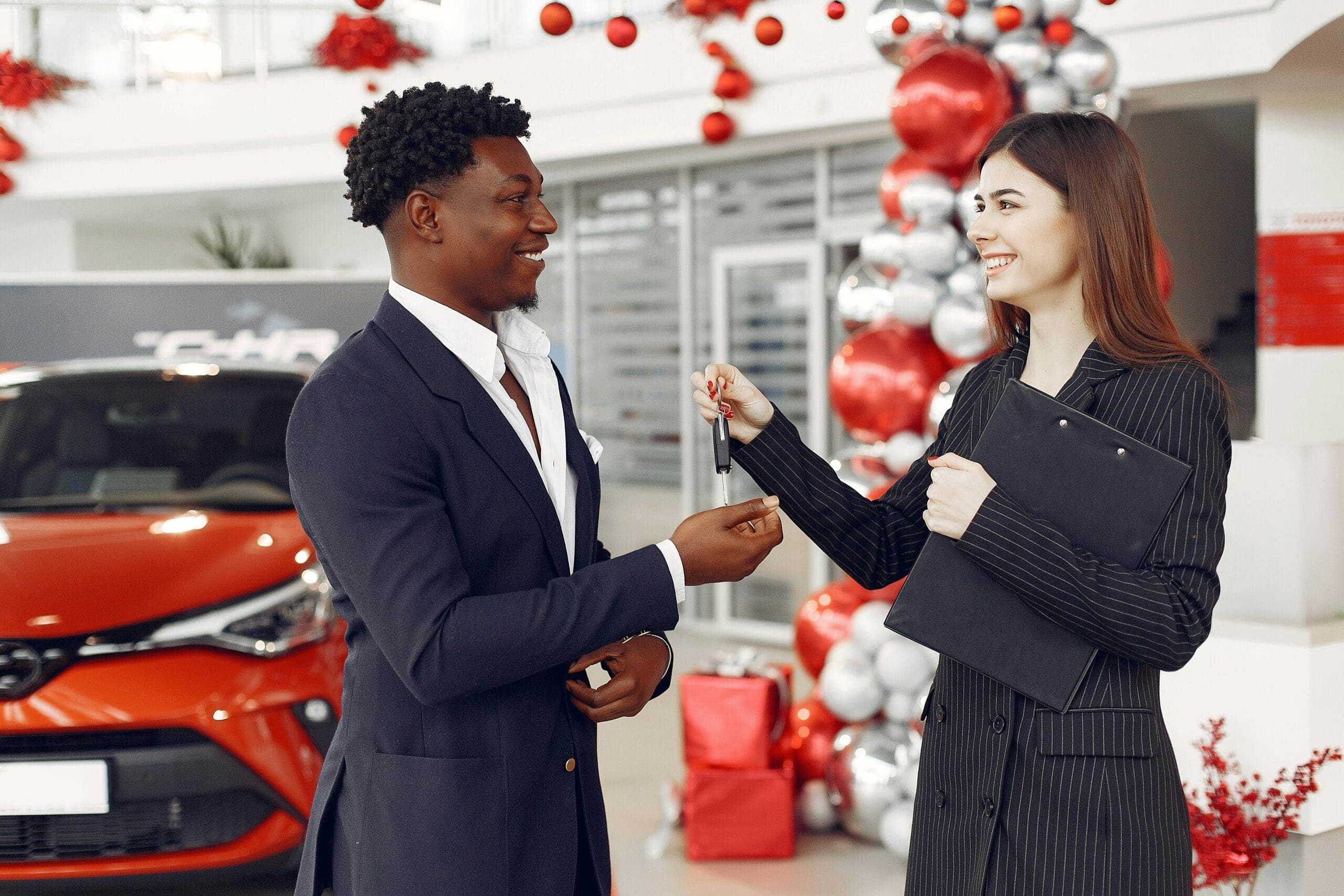 A woman hands over car keys to a man in a car dealership decorated with balloons and ribbons, with a red car in the background. Both individuals are smiling.